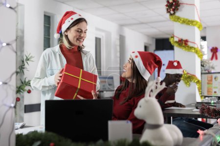 Photo for Thrilled office employee gets Christmas present from asian coworker in festive decorated workspace. Excited worker gets special secret santa gift box from colleague during winter holidays - Royalty Free Image