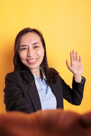 Téléchargez les photos : Joyeuse femme asiatique saluant un ami distant lors d'une réunion par vidéoconférence, souriant lors d'une discussion en ligne. Femme d'affaires satisfaite en costume formel discuter du projet réussi avec l'équipe en studio - en image libre de droit