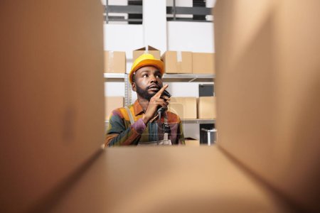 Photo for Selective focus of employee checking cardboard boxes, working at clients orders preparing packages for delivery in warehouse. African american stockroom worker wearing industrial overall and helmet - Royalty Free Image