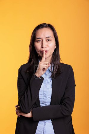 Photo for Portrait of filipino woman making shh gesture in studio by putting forefinger over lips posing over yellow background. Silent adult shushing people gesturing secrecy and confidentiality. - Royalty Free Image