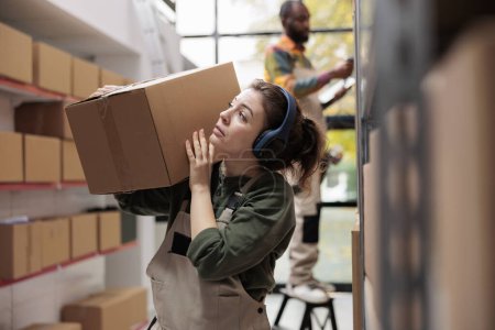 Photo for Woman manager holding cardboard box during inventory, working at customers orders preparing merchandise for delivery. Storage room supervisor wearing headphones listening music in warehouse - Royalty Free Image