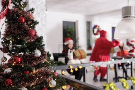 Foto de Árbol de Navidad decorado festivamente de cerca con los trabajadores de negocios que reciben regalos de Santa Claus en un fondo borroso. Navidad adornan el espacio de trabajo moderno durante la temporada de vacaciones de invierno - Imagen libre de derechos
