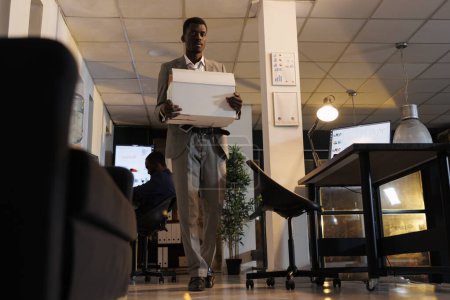 Photo for African american entrepreneur holding cardboard box with his belongings after being fired from business startup office. Advisor agent quitting corporate job late at night. Dismissal concept - Royalty Free Image