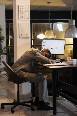 Téléchargez les photos : Gestionnaire exécutif épuisé surmené dormant sur la table de bureau dans le bureau de démarrage après avoir planifié le plan d'investissement de l'entreprise tard dans la nuit. Entrepreneur accro au travail travaillant à un projet de marketing. - en image libre de droit