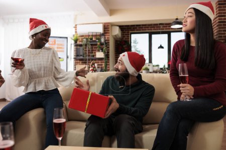 Photo for Excited man with festive wrapped gift box guessing secret santa while celebrating christmas in office. Cheerful coworkers in xmas hats exchanging presents and enjoying new year holiday - Royalty Free Image
