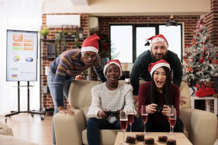Foto de Compañeros de trabajo corporativos que tienen alegre celebración de Navidad, jugando videojuegos juntos en el sofá. Colegas relajándose y sentados en el sofá con mandos en la oficina decorada festiva - Imagen libre de derechos