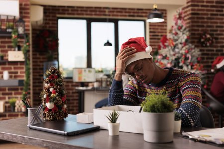 Foto de Deprimido afroamericano hombre en sombrero de santa sentado en el escritorio del lugar de trabajo después de ser despedido en la víspera de Navidad. Empleado molesto de la empresa despedido del trabajo durante la celebración del invierno - Imagen libre de derechos