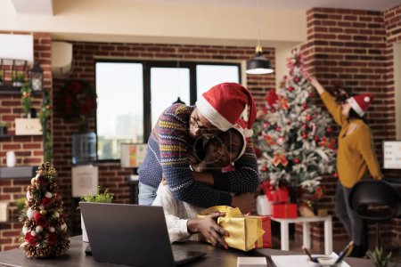 Photo for African american office employee hugging colleague while greeting with christmas gift. Company coworkers embracing while sharing xmas presents and spreading holiday atmosphere - Royalty Free Image