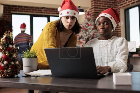 Photo for Company employee sitting at desk and coworking with colleague on project in christmas decorated corporate environment. Diverse women workers in xmas hats doing teamwork during winter holiday - Royalty Free Image
