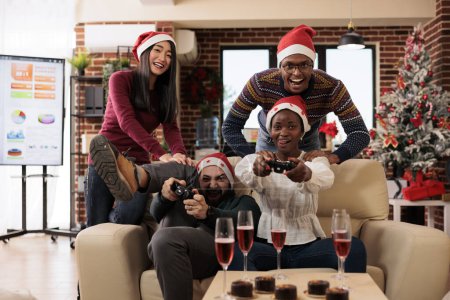 Téléchargez les photos : Une équipe de collègues excités dans santa chapeaux participant à la compétition de jeux vidéo dans un lieu de travail décoré. Travailleurs joyeux célébrant Noël et jouant ensemble au jeu de console - en image libre de droit