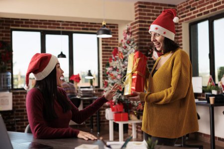 Photo for Excited diverse women coworkes wearing santa hats exchanging xmas gifts while working in decorated office. Happy employee greeting colleague and giving christmas present box - Royalty Free Image