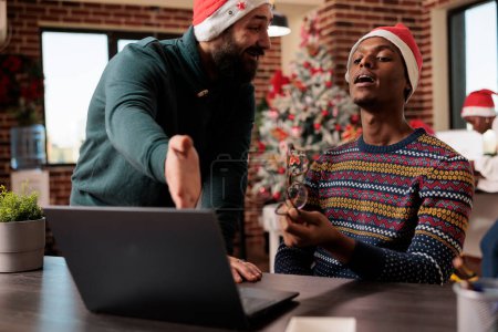 Photo for Company team lead in santa hat helping employee with project while working in christmas decorated office. Manager pointing on laptop and explaining task to coworker in festive workplace - Royalty Free Image