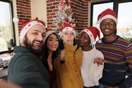 Photo for Smiling diverse men and women colleagues wearing santa hats posing for selfie together in office with christmas decorations. Cheerful coworkers taking group photo at new year corporate party - Royalty Free Image