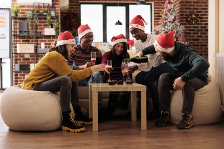 Photo for Cheerful colleagues holding sparkling wine glasses, enjoying festive and merry atmosphere in xmas decorated office. Happy workers team sitting around table and celebrating new year - Royalty Free Image