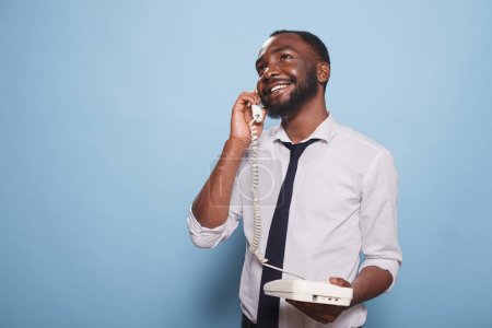 Téléchargez les photos : Homme afro-américain adulte faisant un appel téléphonique sur un téléphone fixe. Homme d'affaires souriant sur un téléphone analogique, bavardant. Un individu contemporain qui s'amuse et prend des appels - en image libre de droit