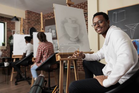 Foto de Feliz chico afroamericano sentado en el caballete en el aula disfrutando de hacer arte durante la clase de dibujo en grupo, espacio creativo para la conexión social. Hombre sonriente aprendiendo a dibujar en el taller de arte - Imagen libre de derechos