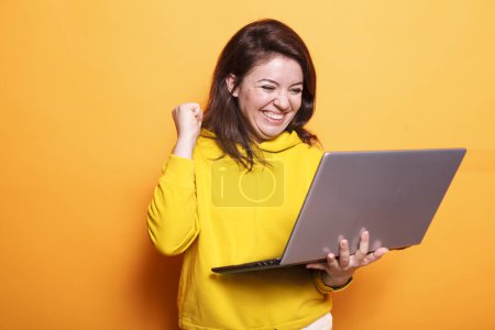 Photo for Portrait of excited lady with her fist in the air while holding personal computer. Beautiful caucasian woman displaying positive emotions after seeing great news on her digital laptop. - Royalty Free Image