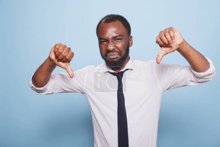 Annoyed black man showing thumbs down gesture on camera, making disagreement and disapproval sign in studio. Displeased individual posing over blue background with negative symbol.