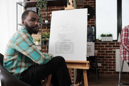 Foto de Hombre afroamericano sentado en el caballete en el aula y mirando a la cámara, estudiando bellas artes. Personas que participan en clases magistrales de dibujo, aprendiendo varias técnicas centradas en el dibujo a lápiz. - Imagen libre de derechos