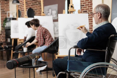 Photo for Senior man with physical disabilities sitting at easel during group art class, wheelchair user increasing fine motor skills through drawing practice. Disabled person engaging in creative activities - Royalty Free Image