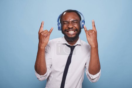 Photo for Enthusiastic young man full of energy doing rock and roll hand sign while listening to music in wireless headphones. African american freelancer making gestures while listening to loud music. - Royalty Free Image
