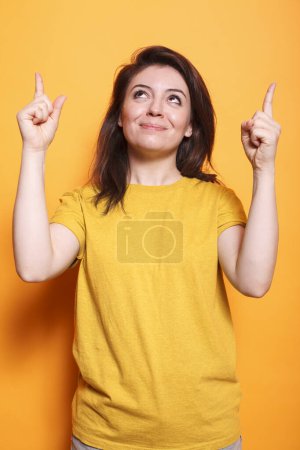 Téléchargez les photos : Portrait de dame brune joyeuse gesticulant avec les index vers le haut et souriant. Femme caucasienne souriante utilisant les mains pour montrer la direction et indiquant vers le haut pour la photo en studio. pose de la publicité. - en image libre de droit