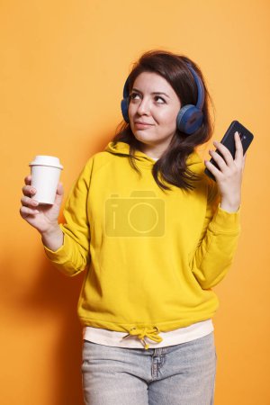 Téléchargez les photos : Portrait d'une femme détendue dans un studio tenant une tasse de café recyclable et écoutant de la musique sur un casque sans fil. Femme individuelle tenant smartphone utilise une application de streaming en ligne. - en image libre de droit