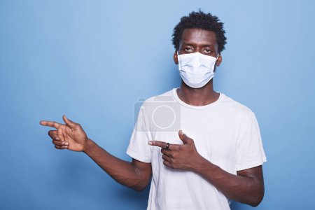 Photo for Afro haired black man in portrait facing the camera and gesturing with his hands to the right. African American guy with a face mask marketing a product while motioning with his arms. - Royalty Free Image