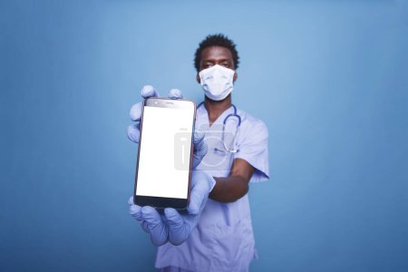 Téléchargez les photos : Homme noir en blouse tenant un téléphone portable avec écran blanc. Gros plan d'un infirmier portant un masque facial et un smartphone avec un gabarit chromatique blanc. - en image libre de droit