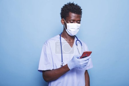 Photo for Black man wearing face mask and gloves browsing for healthcare data through his cell phone. African American male nurse practitioner surfing the net on his digital smartphone. - Royalty Free Image