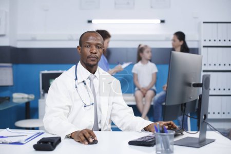Photo for Looking at the camera, African American male doctor sits at desk in hospital office. Portrait of black guy in a lab coat at clinic, with a patient in the background being aided by a nurse. - Royalty Free Image