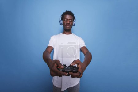 Téléchargez les photos : Joueur vidéo afro-américain en studio utilisant contrôleur et écouteurs. Homme noir jouant à un jeu Internet sur une console avec un joystick tout en se tenant sur fond bleu isolé. - en image libre de droit