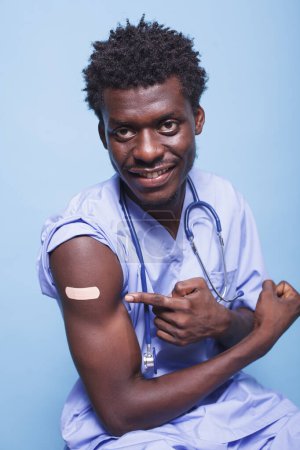 Photo for After coronavirus immunization, African American medical nurse wears adhesive bandage. Male healthcare specialist wears scrubs and stethoscope pointing to vaccination injection plaster for immunity. - Royalty Free Image