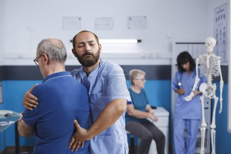 Photo for Orthopedic physician stretches spine and shoulder of senior patient with injury for physiotherapy. Medical practitioner giving help to retired old man with back pain for physical recovery. - Royalty Free Image