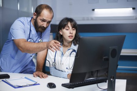 Téléchargez les photos : Infirmière en blouse bleue pointant vers un moniteur PC tout en parlant à un médecin portant un blouse de laboratoire. Les travailleurs médicaux sont vus utilisant un ordinateur et ayant une discussion approfondie sur les soins de santé. - en image libre de droit