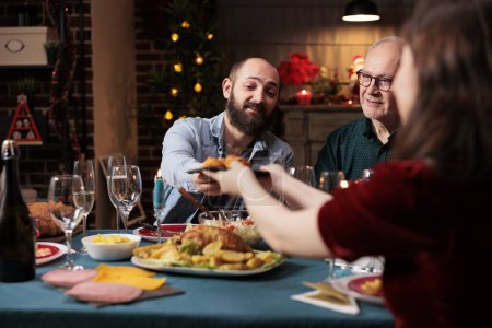 Téléchargez les photos : Les personnes mangeant de la nourriture au dîner festif, célébrant la veille de Noël ensemble et passant des assiettes de repas autour. Groupe joyeux de personnes appréciant l'événement de célébration de vacances à la maison, fête d'hiver. - en image libre de droit