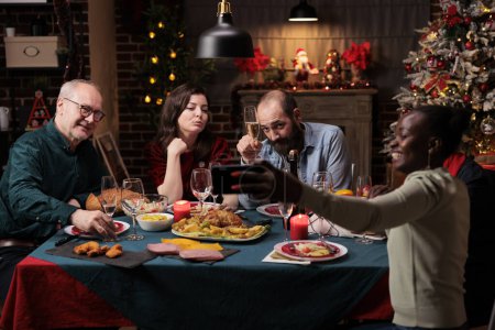 Photo for Persons taking photos at festive dinner, making memories of christmas eve celebration at home. Diverse friends and family having fun with drinks and food, taking pictures with smartphone. - Royalty Free Image
