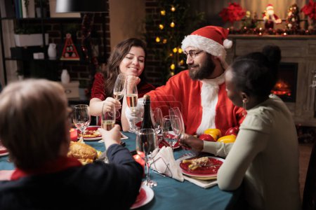 Photo for Young man santa claus having fun with friends at dinner, enjoying christmas holiday spirit together at table. People celebrating seasonal event with food and alcohol, gathering to tell stories. - Royalty Free Image