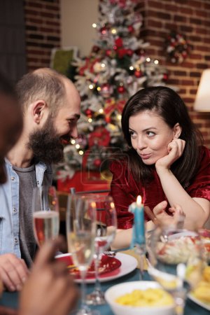Photo for Cute couple enjoying christmas eve dinner with friends, celebrating holiday with traditional food. Diverse people gathering at home with family to eat and drink wine on xmas event. - Royalty Free Image