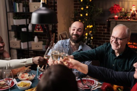 Photo for Diverse people clinking wine glasses at christmas eve dinner, celebrating holiday event with alcohol and homemade food. Friends and family saying cheers with drinks at table, cozy decorations. - Royalty Free Image