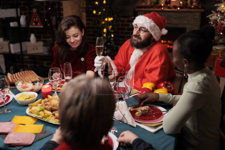 Téléchargez les photos : Adulte en costume santa claus à table avec diverses personnes, profiter de fête la veille de Noël. Jeune en costume et chapeau répandant la positivité pendant la célébration de décembre. - en image libre de droit