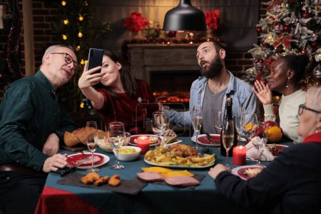 Téléchargez les photos : Les gens prennent des photos lors du dîner festif avec de la nourriture maison et des verres de vin, faisant des souvenirs pendant les vacances d'hiver de la veille de Noël. Diverses personnes s'amusent avec des photos au téléphone à la maison. - en image libre de droit