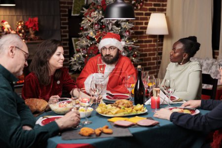 Téléchargez les photos : Heureux santa claus homme faire rire les gens à table, célébrer les vacances de Noël ensemble à la maison. Personnes âgées, amis et famille se réunissant près du Père Noël pour raconter des histoires et s'amuser. - en image libre de droit