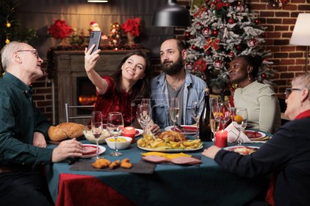 Photo for Happy people takes photos together at home, enjoying dinner feast with glasses of alcohol surrounded by xmas decorations. Diverse friends and family taking pictures during christmas celebration. - Royalty Free Image