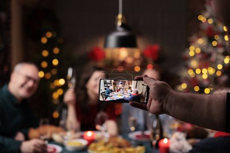Photo for People posing for pictures at home, enjoying festive dinner feast with glasses of wine surrounded by decorations. Diverse persons taking photos during christmas eve holiday celebration. - Royalty Free Image