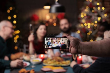 Téléchargez les photos : Homme prenant des photos de famille au dîner, célébrant les vacances de la veille de Noël avec de la nourriture et des boissons traditionnelles. Quelqu'un prend des photos de personnes appréciant la fête avec des verres de vin pendant la célébration. - en image libre de droit