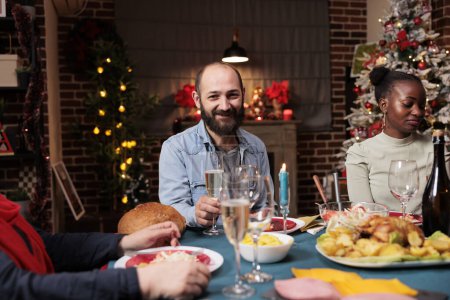 Photo for Young man enjoying festive xmas dinner with diverse people, meeting with family around the table for christmas eve. Diverse persons having fun during winter season celebration, cozy decor. - Royalty Free Image