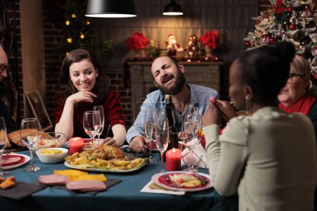 Foto de Gente alegre disfrutando de la fiesta de invierno en la mesa, jóvenes y adultos mayores divirtiéndose comiendo comidas caseras y bebiendo vino. Amigos y familiares se reúnen alrededor para la cena de Navidad festiva. - Imagen libre de derechos