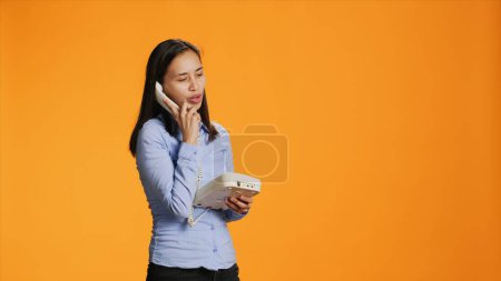 Photo for Asian woman using landline phone for remote call, talking to someone on vintage telephone line. Confident person answering office phone with cord, communicating remotely in studio. - Royalty Free Image
