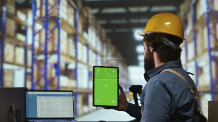 Photo for Warehouse operator holds greenscreen on tablet before working on merchandise distribution, examining isolated display on mobile device. Industrial staff member looks at blank chromakey screen. - Royalty Free Image